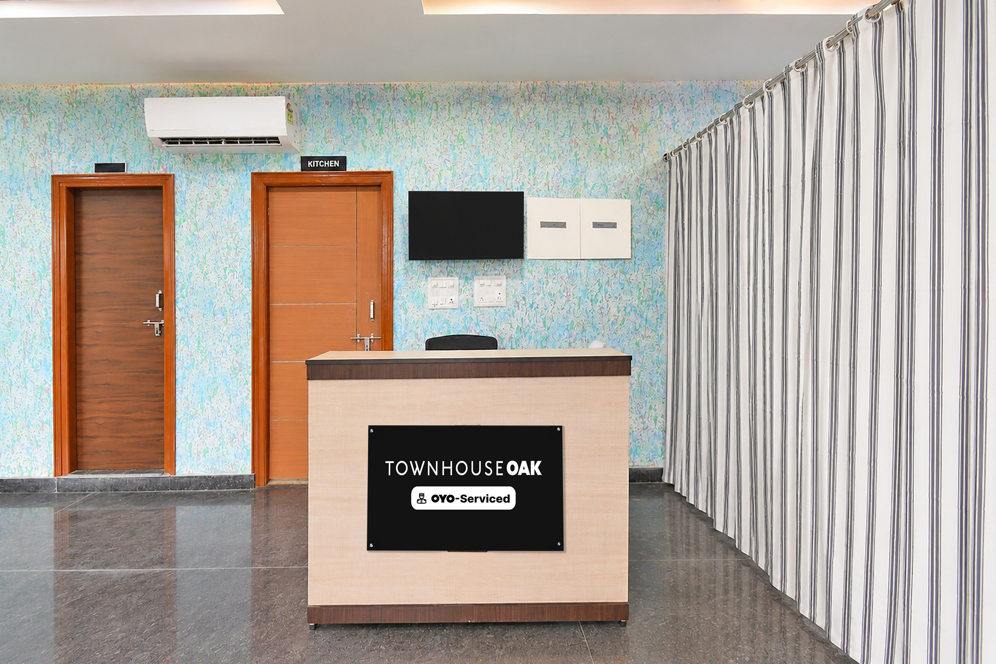En receptionsdisk med en skylt som visar ”TOWNHOUSE OAK” och information om de tjänster som erbjuds av ”OYO-Serviced”. Det finns två dörrar, en väggmonterad tv och en vägg med ett mönster.