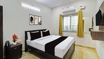 Ein Hotelzimmer mit Bett, Nachttisch, Wandklimaanlage, Vorhängen an einem Fenster und gerahmten Kunstwerken an der Wand.