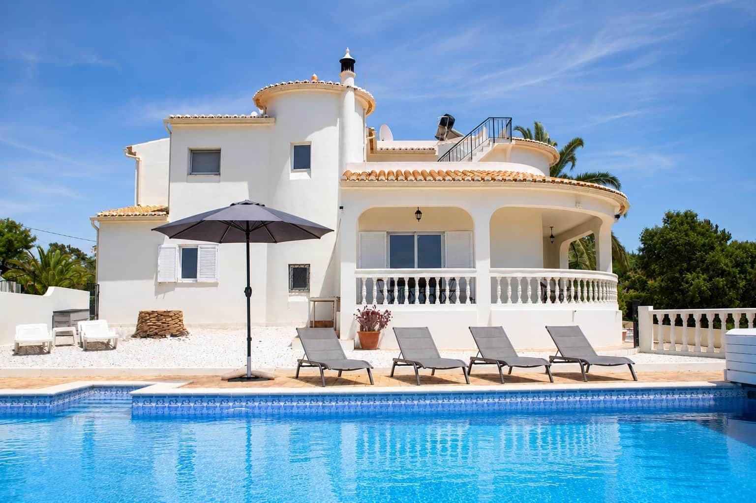 A two-story building with a swimming pool, white walls, and a balcony with sun loungers.