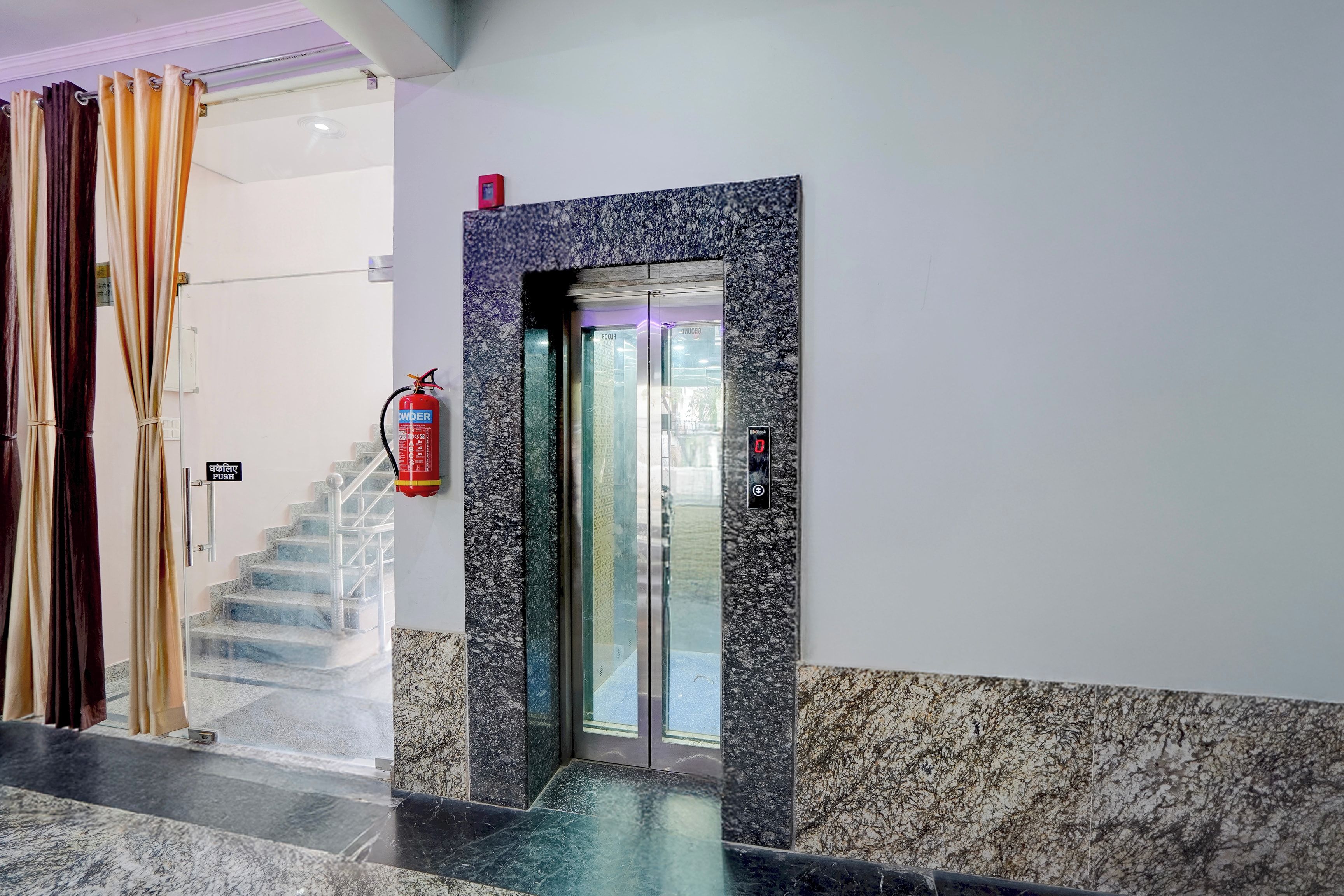 Eine Hotelhalle mit Marmorboden, einem Feuerlöscher, einer Treppe und einem Glastreppenaufzug.