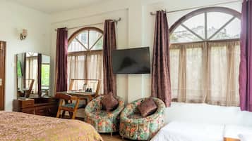 Una camera d'albergo con un letto, due poltrone floreali, una scrivania con uno specchio, un televisore e ampie finestre con tende.
