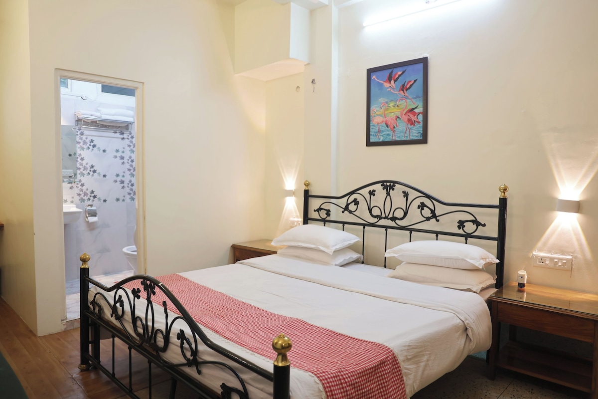 Habitación de hotel con cama, lámparas de noche, un cuadro y un baño visible a través de una puerta de vidrio.