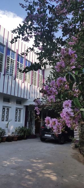 Un edificio con fachada blanca y líneas verticales rosas, un coche estacionado y plantas con flores rosadas en primer plano.