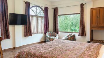 Un dormitorio con una cama grande, televisión, ventana con cortinas, una silla y un armario.