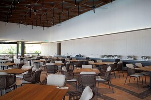 Un amplio comedor interior con numerosas mesas y sillas, una barra larga con estaciones de servicio de alimentos, y un techo con vigas y luminarias a la vista.