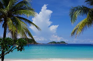 Una playa con agua turquesa cristalina, palmeras y una isla distante.