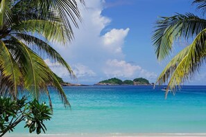 Una playa con agua turquesa cristalina, palmeras y una isla distante.