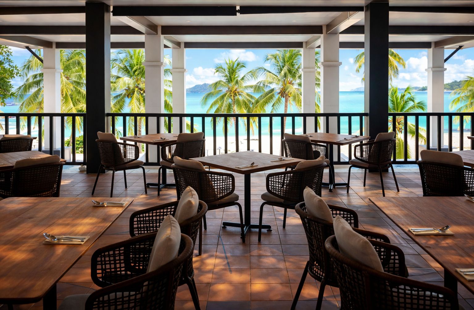 A covered outdoor dining area with tables and chairs, overlooking a beach with palm trees and the ocean.