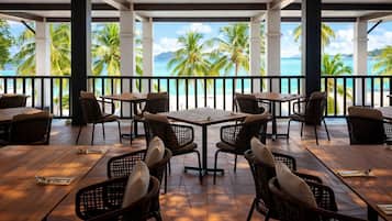 A covered outdoor dining area with tables and chairs, overlooking a beach with palm trees and the ocean.
