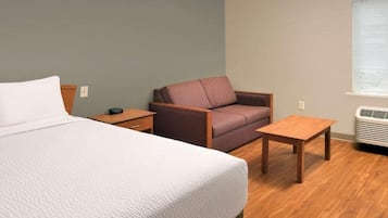 A hotel room with a bed, a chair, a small table, and a window with blinds.