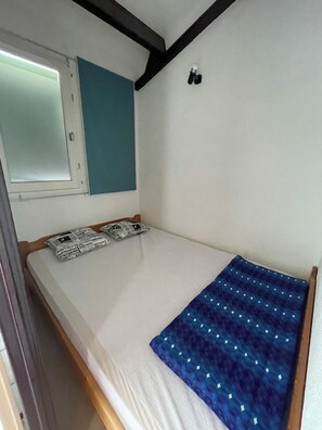 A single bed with a blue patterned bedspread, a window with a view, and a wall-mounted light fixture. - Apartment with a view of the salt flats (Gruissan)
