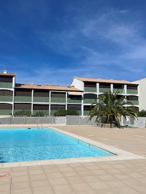 A swimming pool with a tiled deck area, surrounded by a white fence, and a building with multiple balconies and a tiled roof in the background. - Apartment with a view of the salt flats (Gruissan)