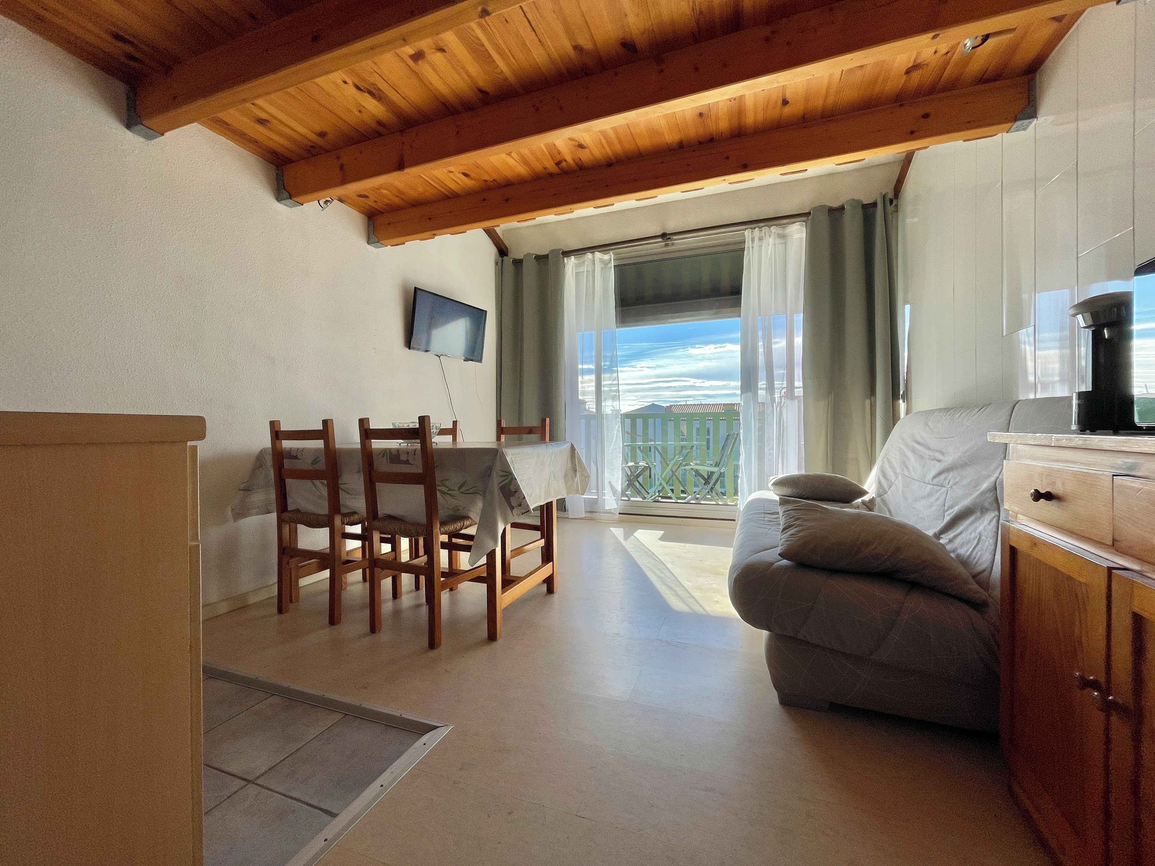 A living room with a wooden ceiling, a sofa, a dining table with chairs, a TV mounted on the wall, and a balcony with a view.