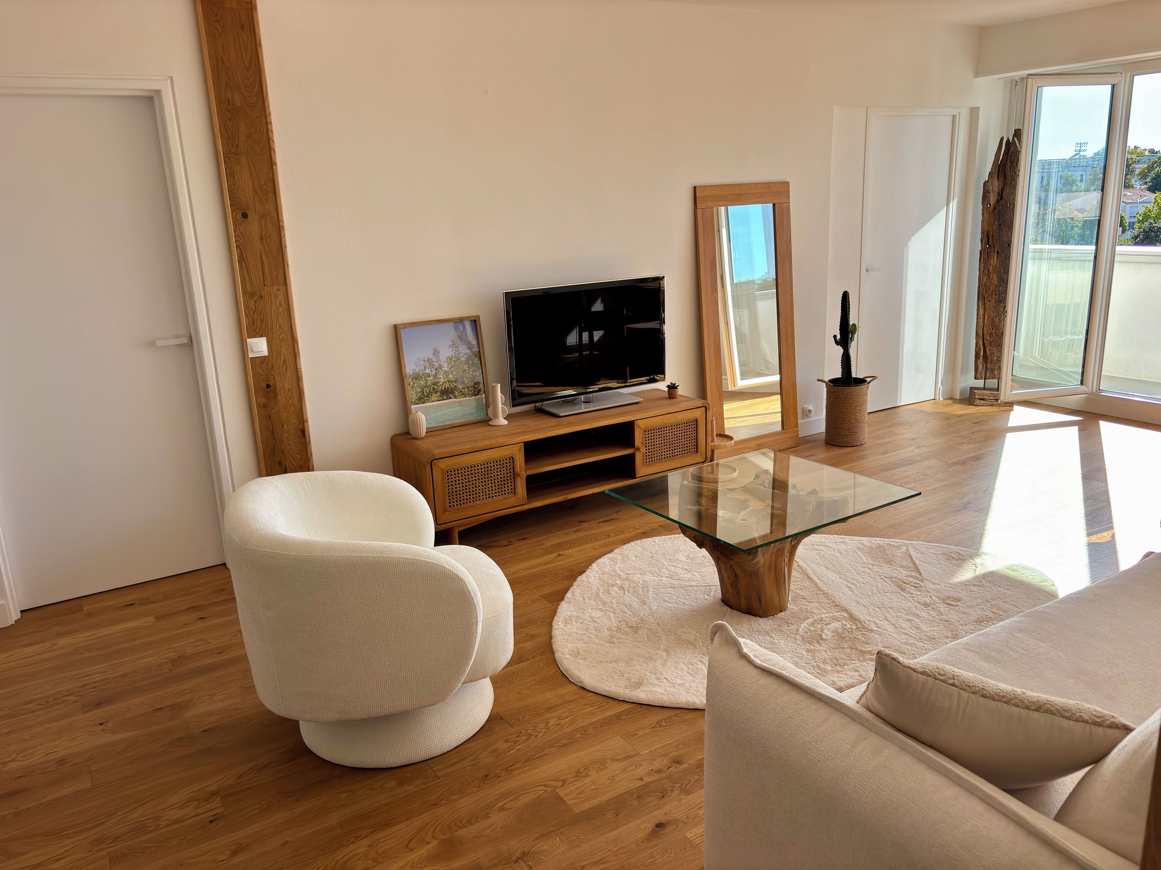 A modern living room with a glass coffee table, white sofas, a wooden TV stand, and a large window with a view.