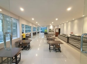 Un amplio comedor de hotel con varias mesas y sillas, una recepción y grandes ventanales que permiten la entrada de luz natural.