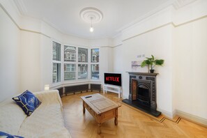 A living room with a fireplace, a television displaying 'NETFLIX', and a large window with blinds. - Willesden Luxe House with Garden (London)