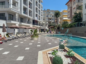 A poolside area with lounge chairs and umbrellas, surrounded by multi-story buildings with balconies. - Qoople Elize C33 - Cozy 1-bedroom apartment in charming Alanya (Alanya)
