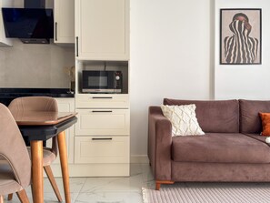 A modern living room with a brown sofa, a dining table with chairs, a kitchen area with cabinets and a microwave, and a framed artwork on the wall. - Qoople Elize C33 - Cozy 1-bedroom apartment in charming Alanya (Alanya)