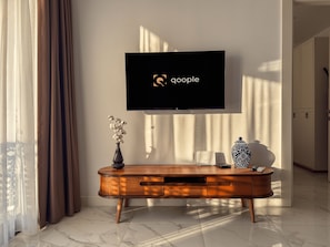 A modern living room with a flat-screen TV mounted on the wall displaying the Google logo, a wooden console table with a vase of flowers, and a decorative ceramic piece. - Qoople Elize C33 - Cozy 1-bedroom apartment in charming Alanya (Alanya)