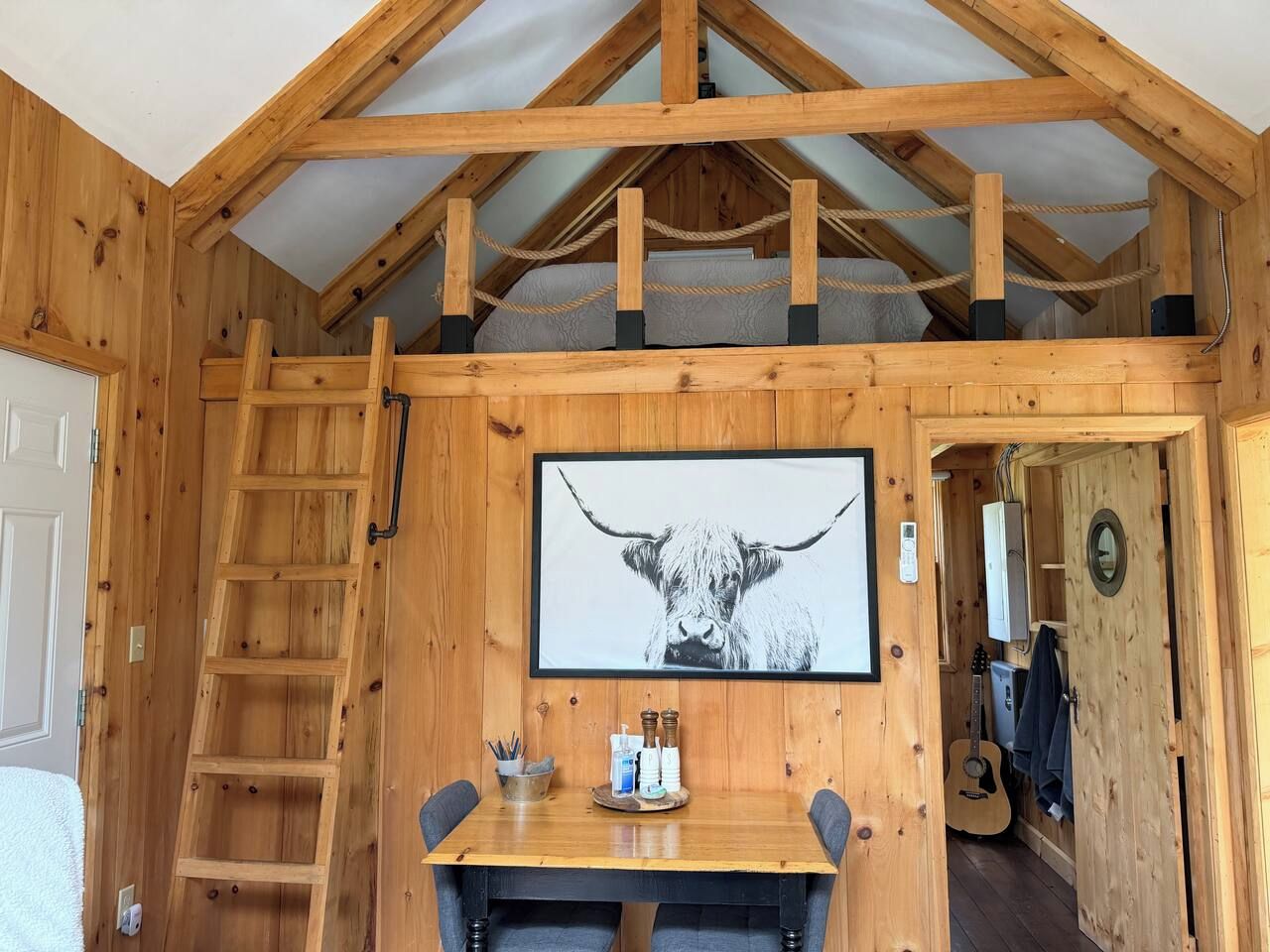 A wooden interior with a lofted ceiling, a framed artwork of a bull, a ladder, and a dining area with a table and chairs.