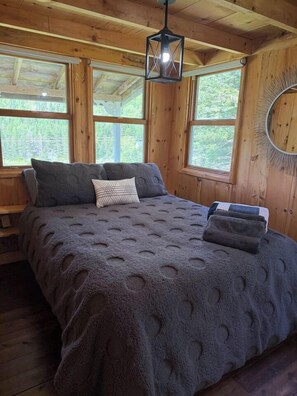 A wooden cabin bedroom with a bed, pillows, a hanging light fixture, and a window with a view of greenery.