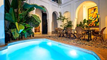 Un área de piscina con zona de comedor y plantas tropicales.