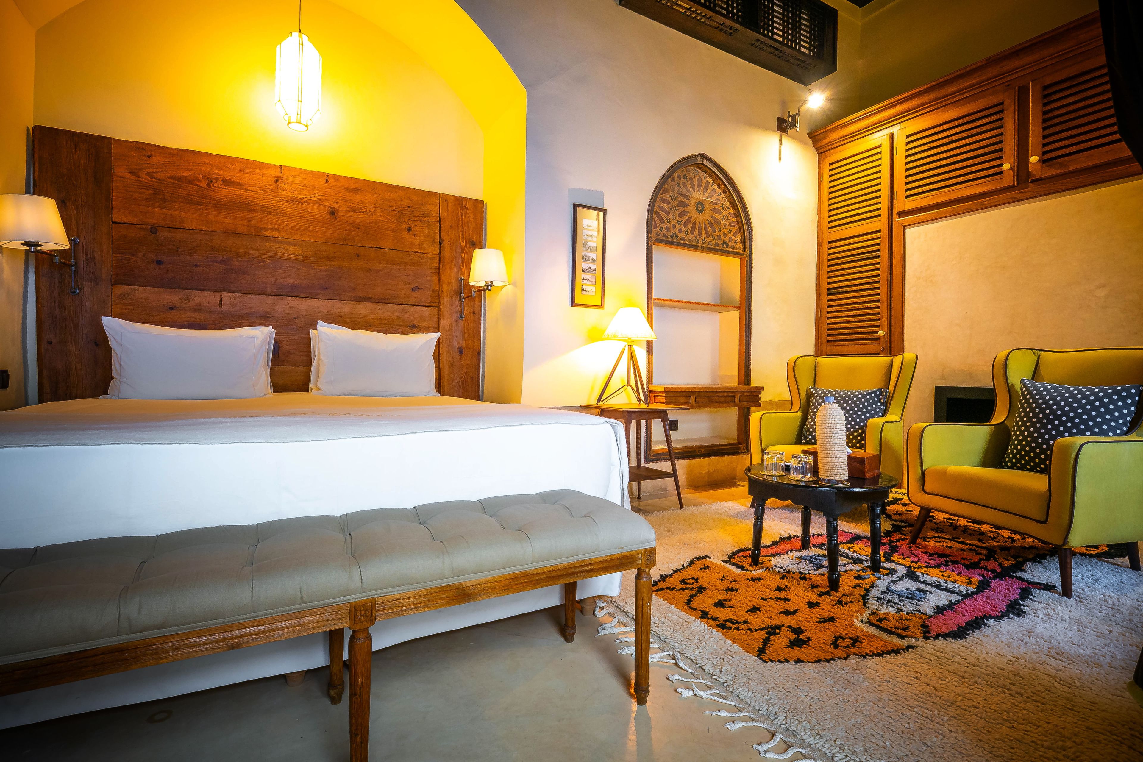 Habitación de hotel con una cama grande, un banco, una mesita, una silla y un espejo decorativo.