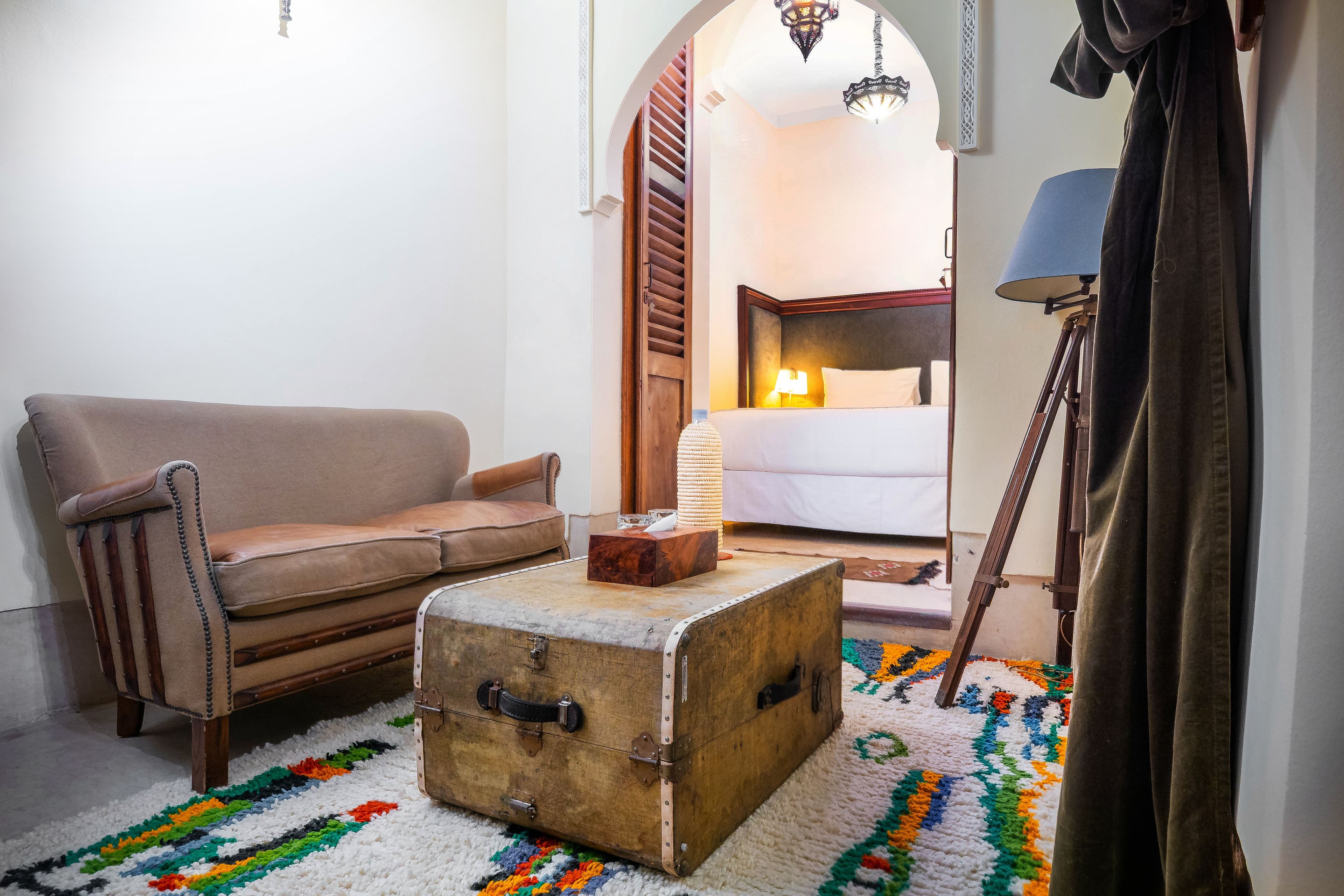 Una habitación acogedora con un sofá de cuero marrón, una maleta antigua, una alfombra colorida, una lámpara y una cama con una mesita de noche.