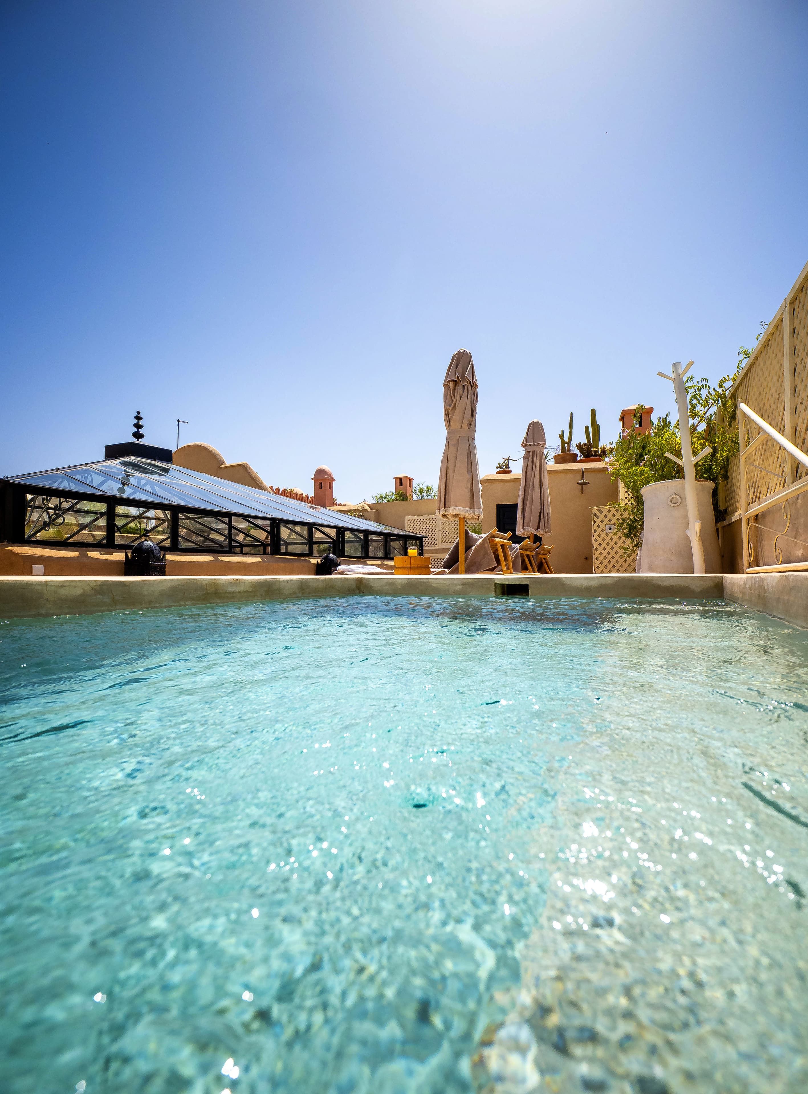 Piscina límpida com teto de vidro, rodeada por espreguiçadeiras e guarda-sóis, e céu azul acima.