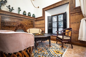 Quarto tradicional com cabeceira de madeira entalhada, uma mesa redonda e um tapete estampado, além de uma janela com grades de ferro decorativas.
