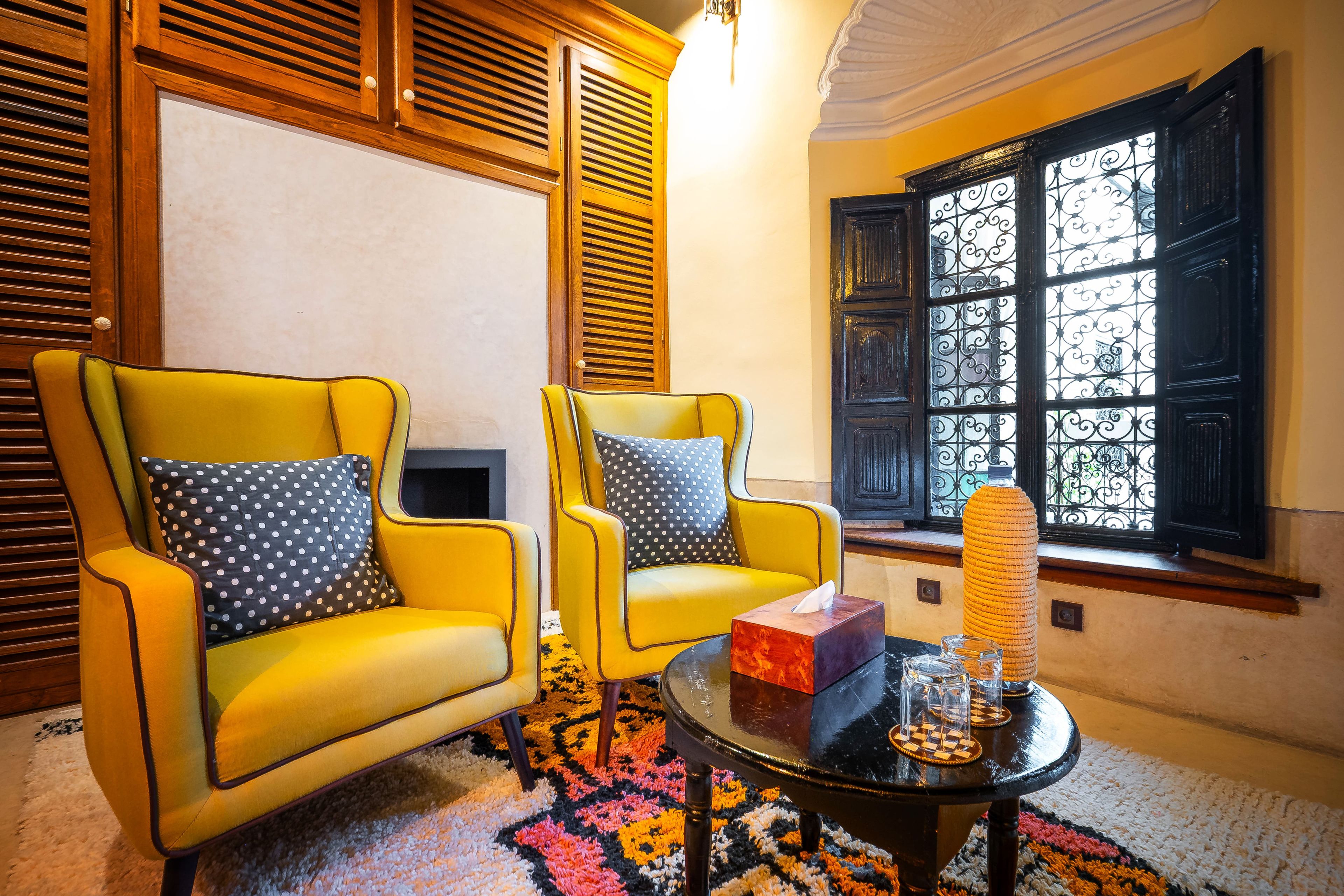 Quarto com duas poltronas amarelas, uma mesa de centro preta e uma janela com grades de ferro decorativas.