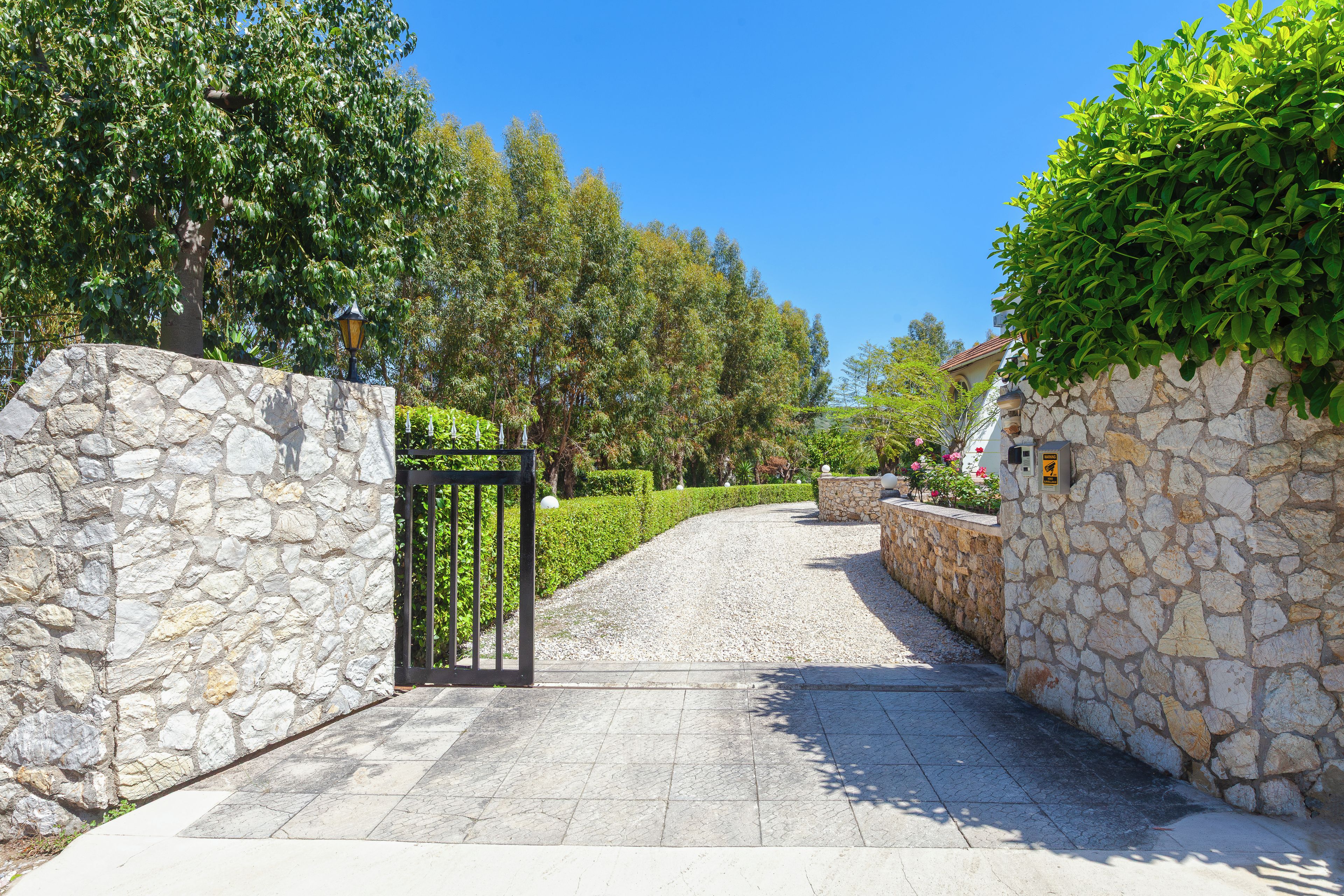 A stone wall entrance with a metal gate, leading to a paved pathway flanked by greenery and a clear blue sky.