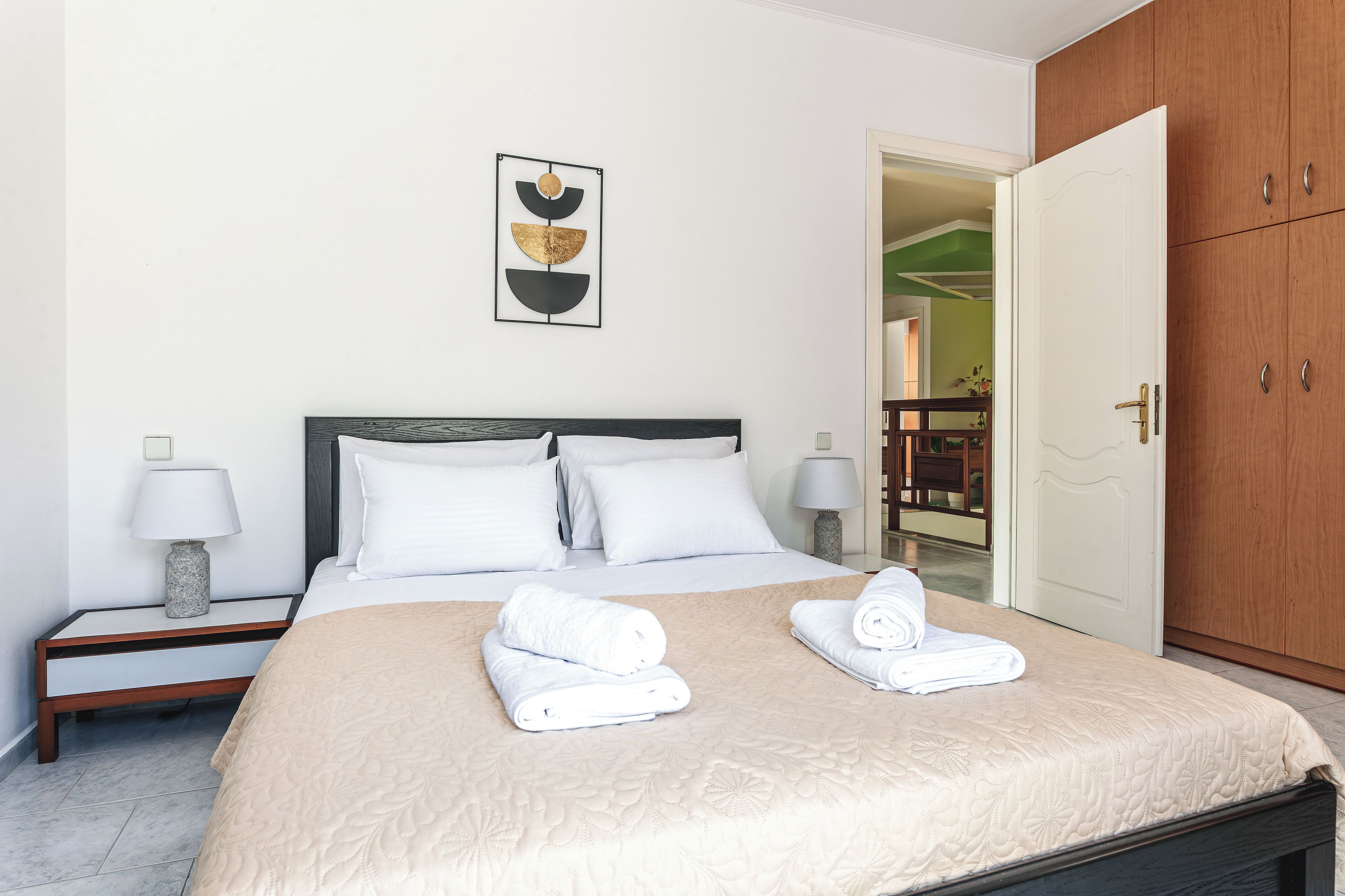 A hotel room with a bed, two bedside tables, a nightstand, a wooden wardrobe, and a framed artwork on the wall.