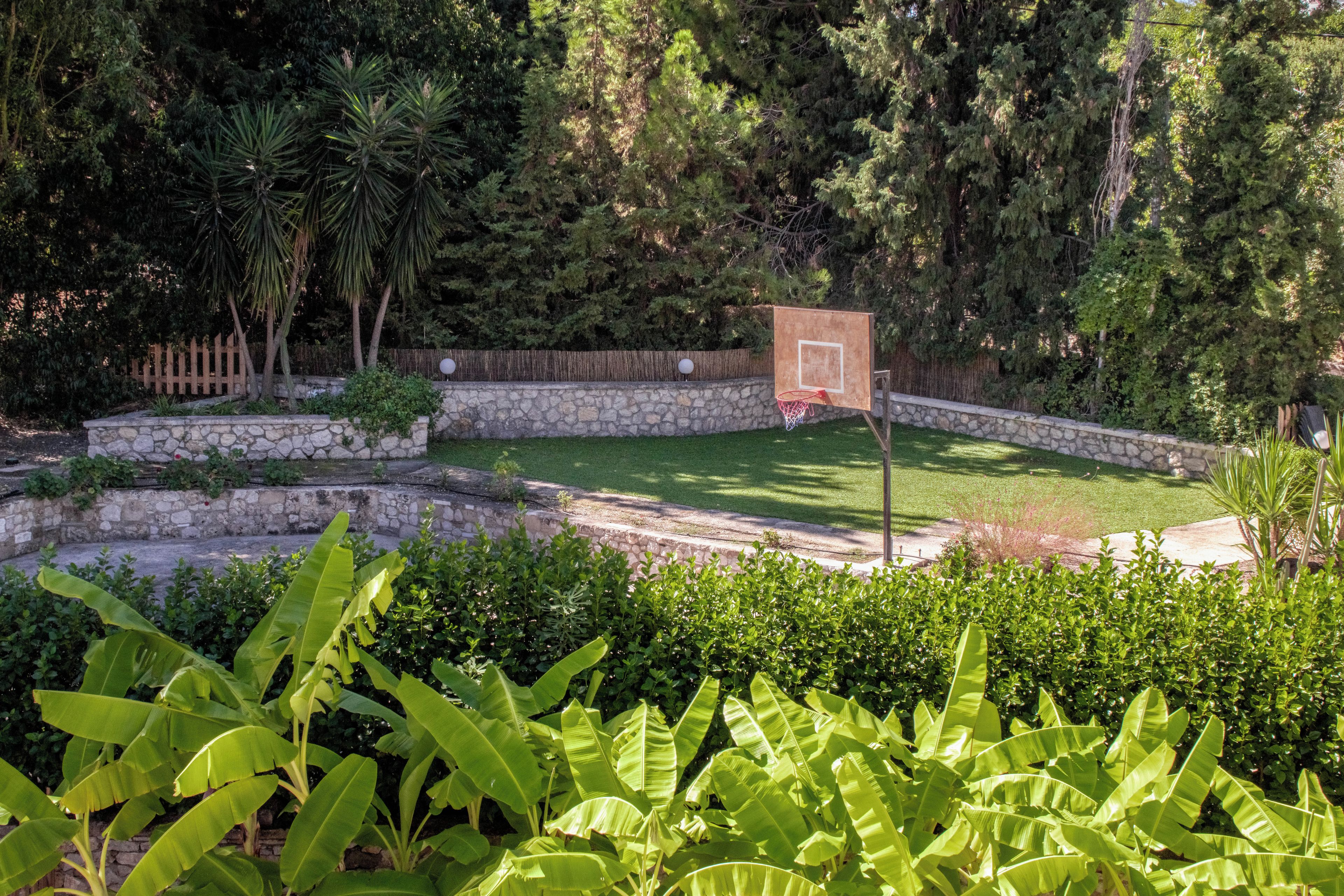 A basketball court with a hoop, surrounded by a stone wall and lush greenery.