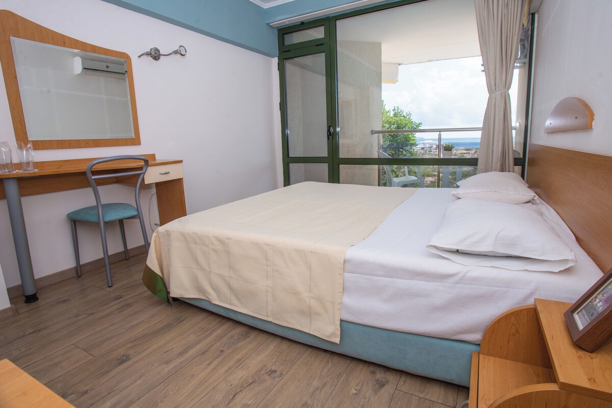 Habitación de hotel con cama, escritorio, silla, espejo y vista al exterior.