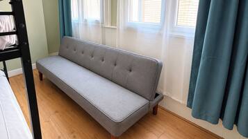 A room with a grey sofa, wooden flooring, a bunk bed, and a window with curtains.