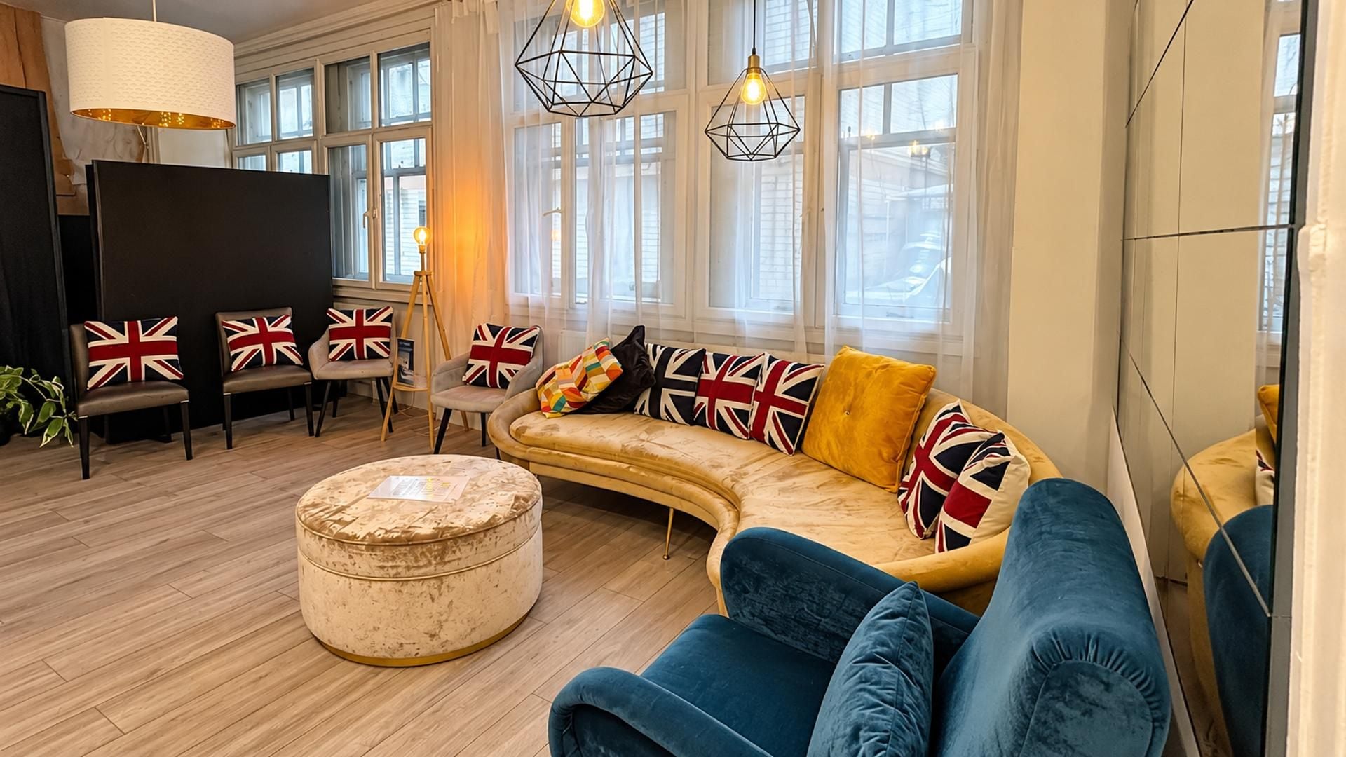 A modern living room with a curved sofa, a round ottoman, and a flat-screen TV. There are multiple chairs with British flag cushions, and the room has large windows with natural light.