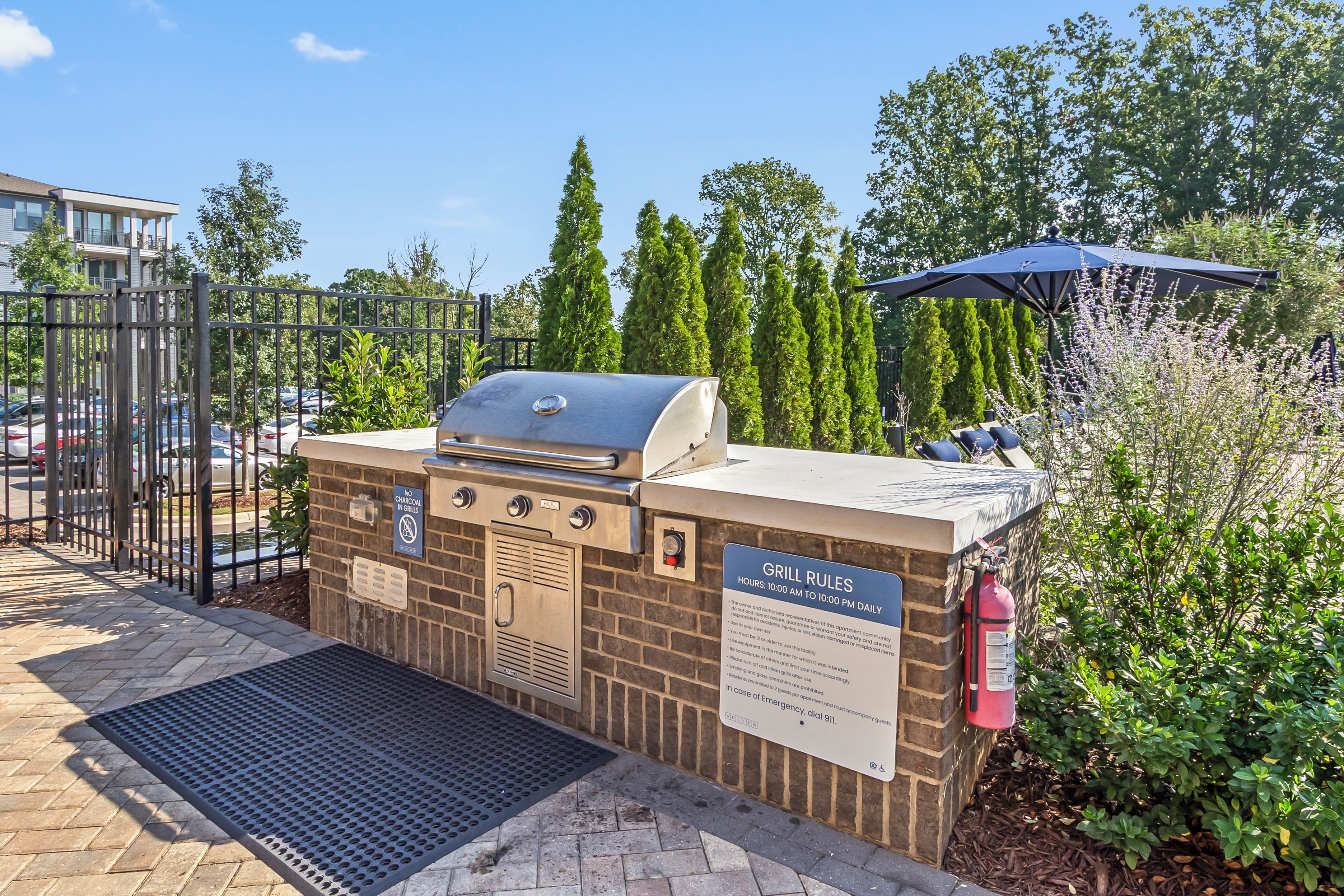 Ett utomhusområde med en grillplats byggd av tegelstenar, komplett med en grill av metall, en brandsläckare och en skylt som beskriver reglerna för grillplatsen.