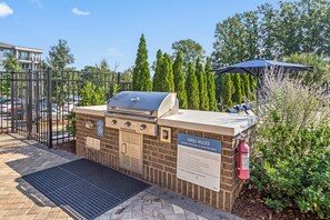 Ett utomhusområde med en grillplats byggd av tegelstenar, komplett med en grill av metall, en brandsläckare och en skylt som beskriver reglerna för grillplatsen.