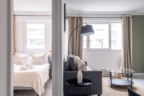 Quarto de hotel com cama, sofá, mesinha, abajur e vista para a cidade pela janela.