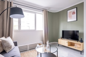 Una sala de estar moderna con un sofá, una mesa de centro de vidrio, un televisor sobre un mueble de madera y un ventanal con cortinas.