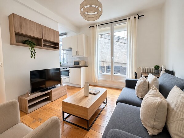 Un salon moderne avec un téléviseur à écran plat, une table basse en bois, un canapé gris et une cuisine en arrière-plan.