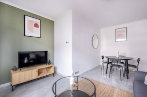 Una sala de estar moderna con un televisor de pantalla plana, un mueble de entretenimiento de madera, una mesa de centro de vidrio, una zona de comedor con una mesa redonda y sillas, y un sofá gris.