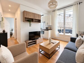 A modern living room with a sofa, coffee table, and TV, a kitchen area with cabinets and appliances, and a large sliding glass door to a balcony. - Magnifique Appartement Levallois Perret (Levallois-Perret)