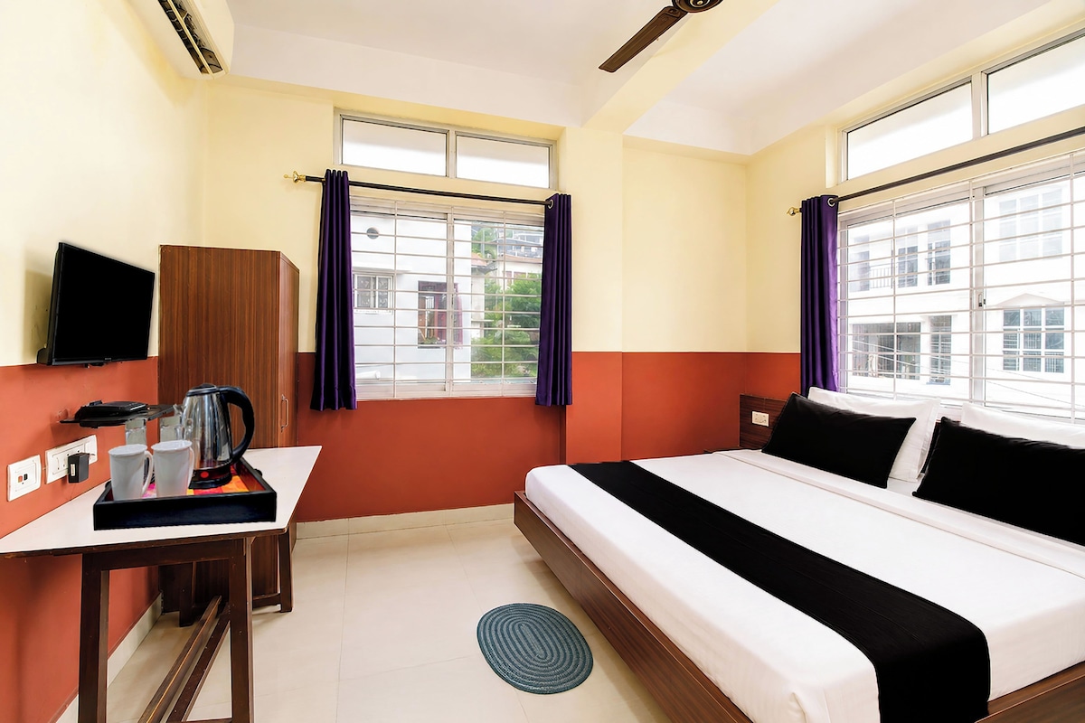 A hotel room with a bed, a desk with a kettle, a TV, and a window with curtains.