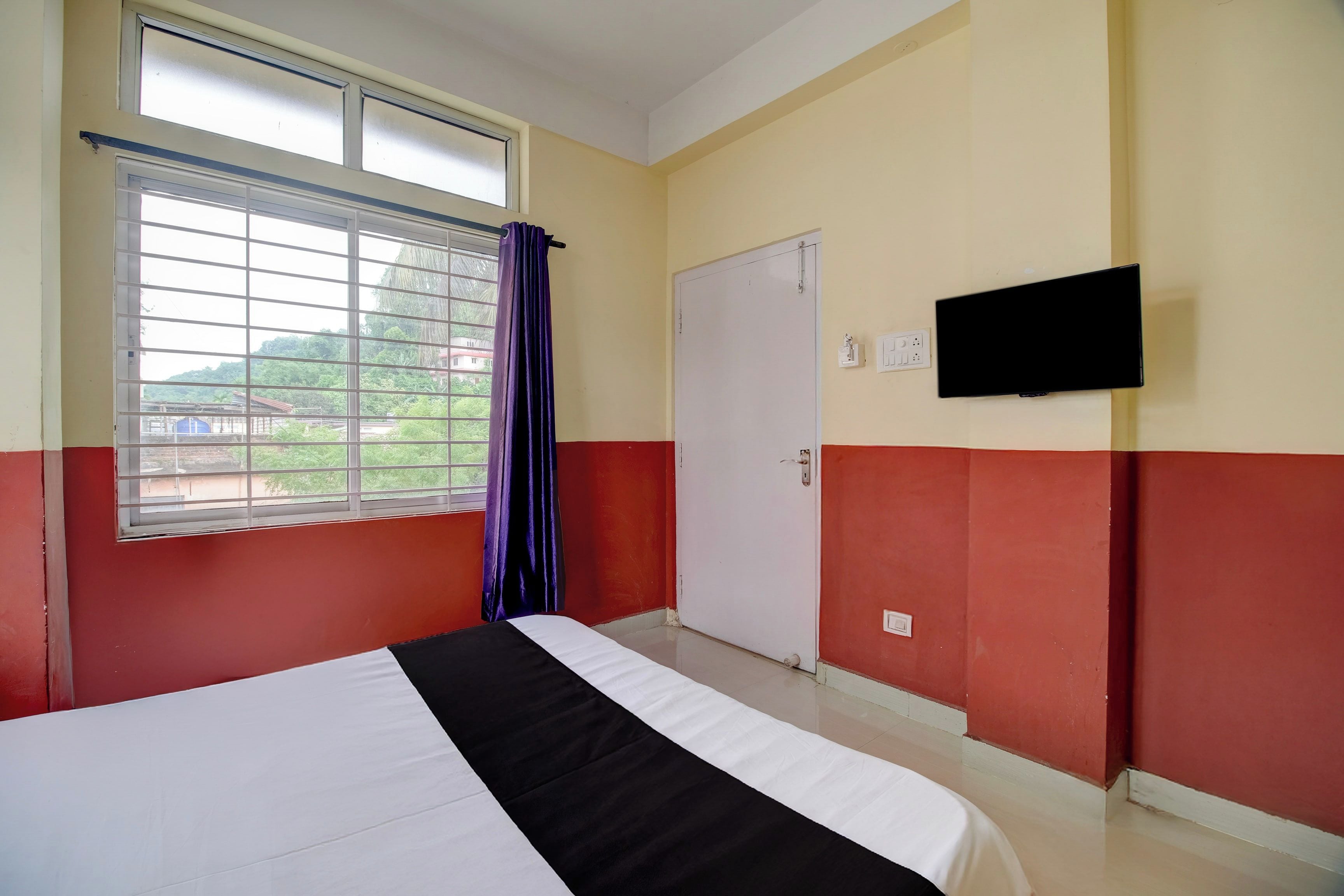 Habitación de hotel con cama, ventana con cortinas, puerta y televisor de pantalla plana montado en la pared.
