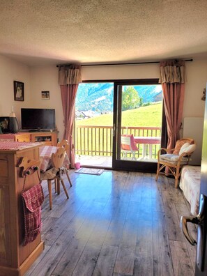 Ein Zimmer mit Holzboden, Fernseher, Esstisch und Blick auf eine grüne Landschaft durch eine Schiebetür.