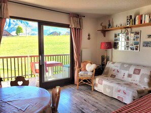 A cozy room with a wooden floor, a sofa, a chair, a table, and a balcony with a view of a green field. - Mountain Apartment for 4 People (Albiez-Montrond)