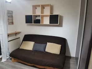A room with a brown sofa, two pillows, a wooden shelf unit, and a radiator. - Cozy Studio in Morillon (Morillon)