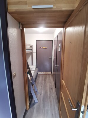 Een smalle gang met een stapelbed, een deur met kapstok en een houten plafond.